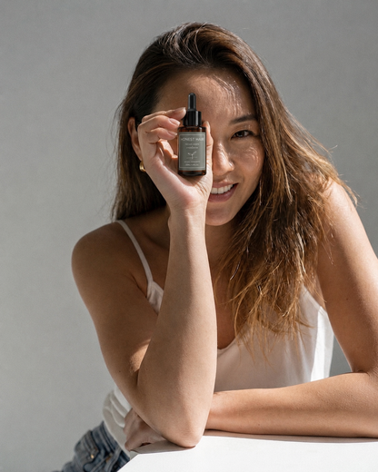 A woman in a white camisole holds a small amber hair serum dropper bottle in front of her face showing satisfaction with the product 