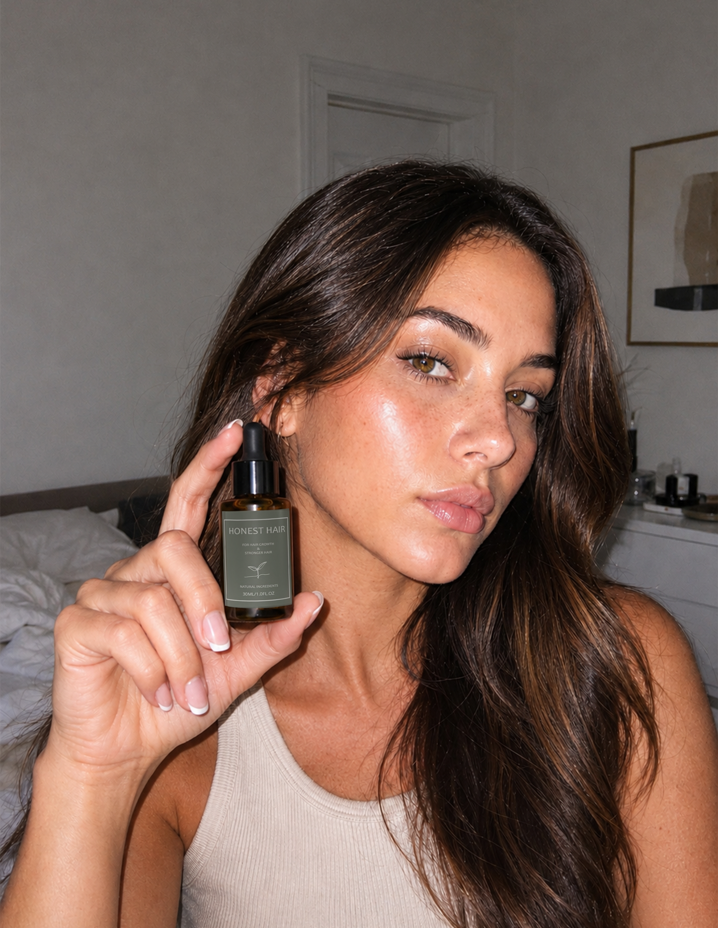 A woman with long dark brown hair holds up a small amber glass dropper bottle with a black cap. The Hair Serum bottle has a dark olive-green label 