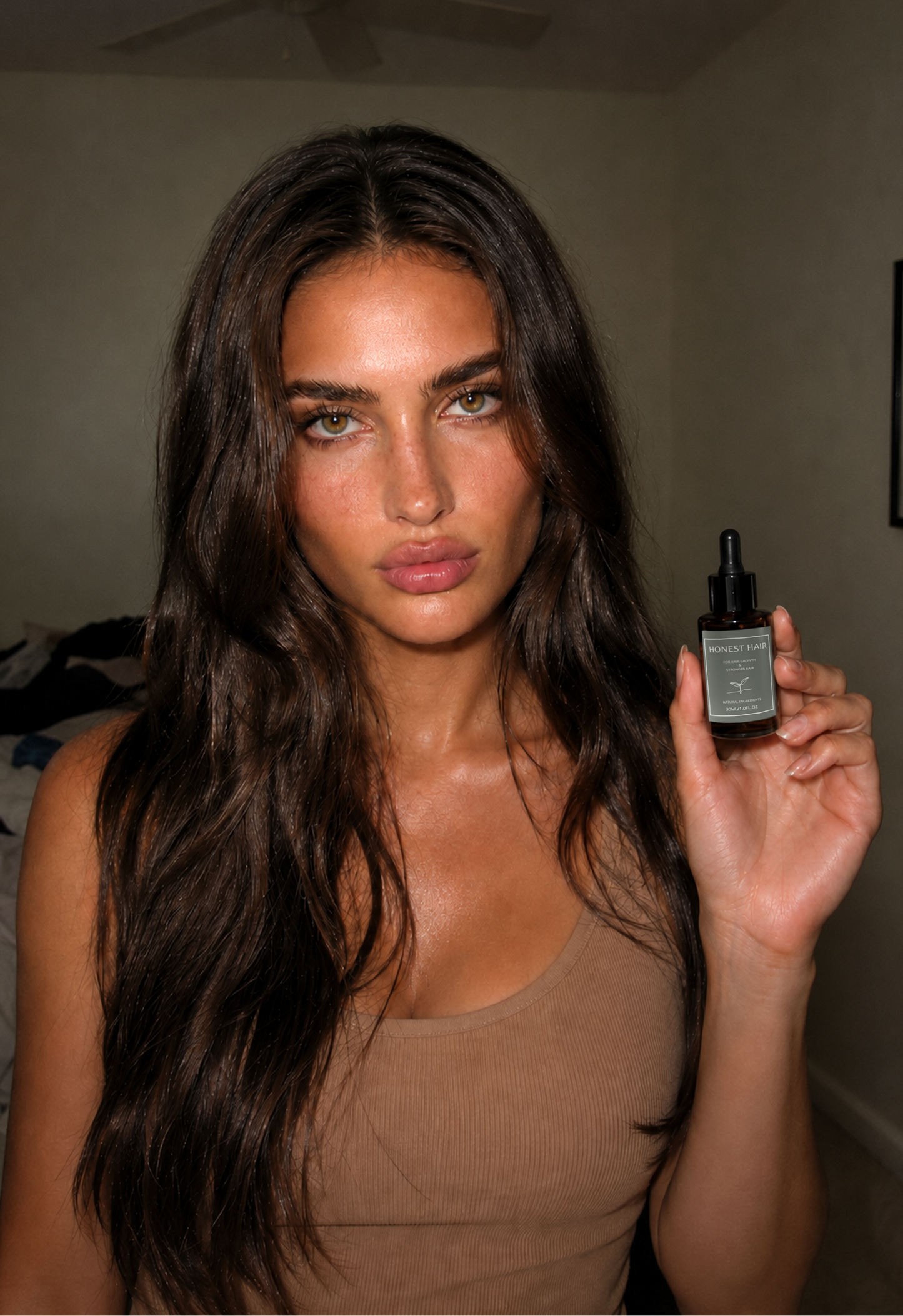 A woman with long, dark brown hair holds up a small hair serum dropper bottle in a bedroom setting.
