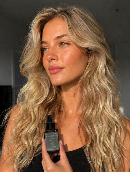 A woman with long, wavy blonde hair holds a dark bottle labeled 'HONEST HAIR' serum  in front of her face. The background is a dimly lit, neutral indoor setting.