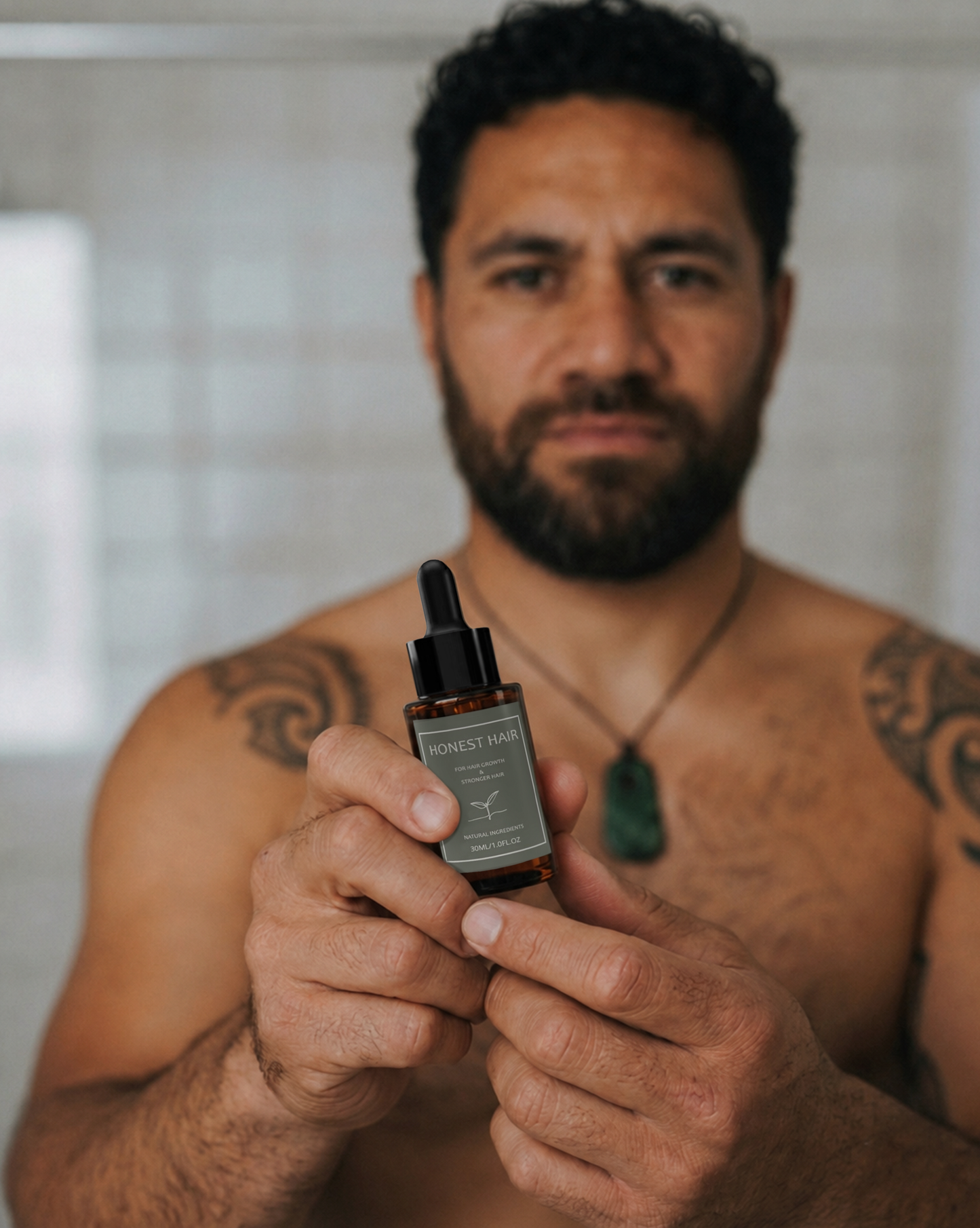 A man with tattoos and a beard holds a small hair serum amber dropper bottle  labeled Hones Hair in front of his face, with a softly blurred bathroom-like background.